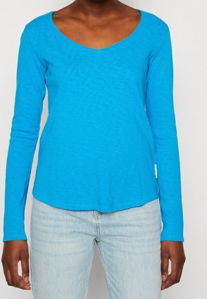 Langærmet V-hals top i lys blå, med et blødt, tekstureret stof og en rund kant, parret med lyse jeans.