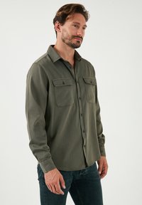Olijfgroene blouse met lange mouwen, twee borstzakken, knoopsluiting aan de voorkant, ronde zoom en een gladde stofstructuur. Gecombineerd met donkere jeans.