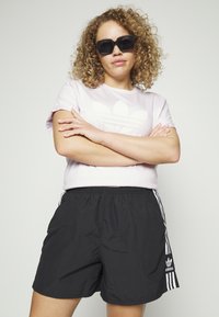 T-shirt rosa chiaro con logo Adidas bianco, abbinata a pantaloni corti neri con righe laterali bianche e cintura elastica. Occhiali da sole neri.