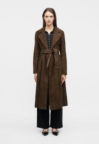 Femme portant un trench-coat en daim marron ceinturé, par-dessus un haut noir boutonné et un pantalon large, avec des chaussures noires à bout pointu, debout devant un fond uni.