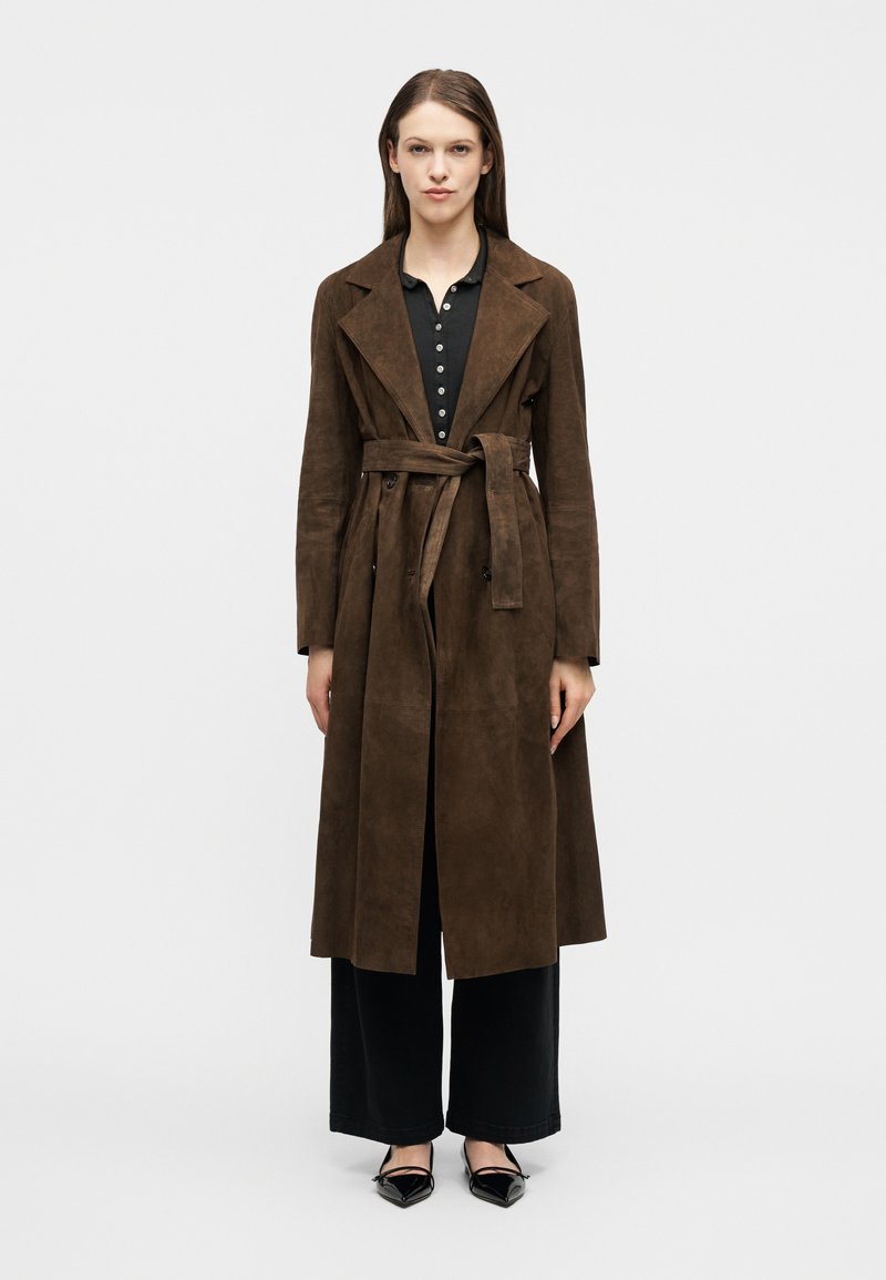Femme portant un trench-coat en daim marron ceinturé, par-dessus un haut noir boutonné et un pantalon large, avec des chaussures noires à bout pointu, debout devant un fond uni.