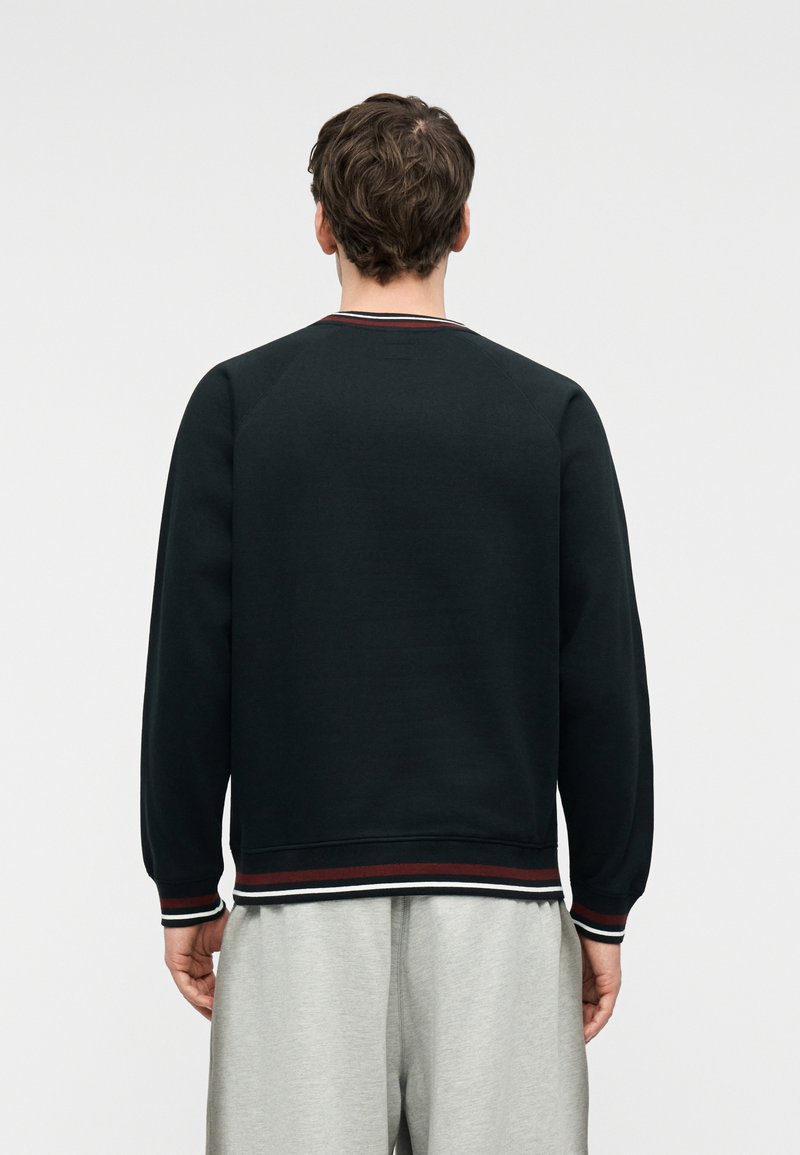 Man wearing a black sweatshirt with red and white striped cuffs and hem, facing away, paired with light gray pants.