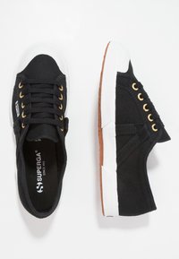 Schwarze Leinen-Sneaker mit goldenen Ösen, weißer Gummikappe und brauner Gummisohle. Sie verfügen über ein niedriges Design und eine Schnürung.