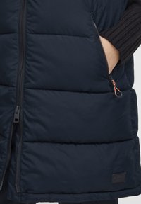 Marinblå puffer-väst med dragkedja fram och fickor, med en mjuk textur. En anmärkningsvärd orange detalj på dragkedjans draghandtag.