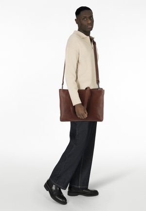 Borsa tote in pelle marrone con manico superiore e tracolla regolabile, texture liscia e design minimalista. La borsa è tenuta da una persona.