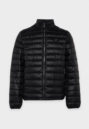 Un homme porte une veste noire matelassée, un pull à motifs, un pantalon noir, une casquette et des chaussures massives sur un fond blanc uni.