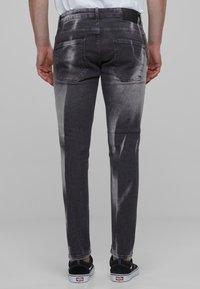 Slim-fit pelēki džinsi ar izbalējušu dizainu, kas apveltīti ar divām aizmugurējām kabatām un ādas uzlīmi aizmugurē ap vidukli. Tekstūra izskatās gluda.