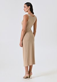 Robe beige cintrée, longueur genou, avec un dos en décolleté et un éclat métallique, associée à des sandales dorées à lanières. Texture lisse.