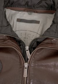 Braune Lederjacke mit einer schwarzen Nylonkapuze, ausgestattet mit zwei Metallreißverschlüssen und einem gesteppte Innenfutter. Das innere Etikett ist vor einem hellen Hintergrund sichtbar.