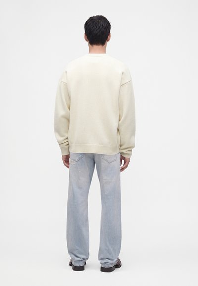 Homme debout, de dos, portant un pull crème, un jean bleu clair à jambes larges et des chaussures noires sur un fond blanc uni.