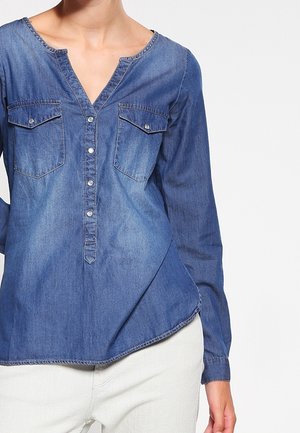 Femme portant une chemise en denim bleu à manches longues avec des poches poitrine boutonnées et un pantalon blanc, sur un fond clair uni.
