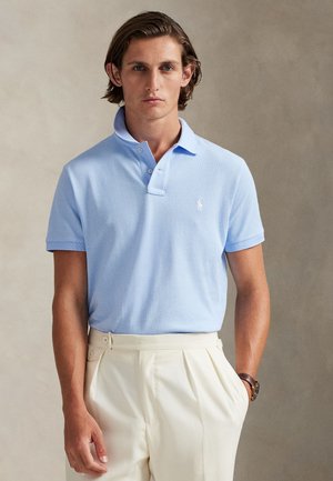 Homme portant un polo bleu clair à manches courtes rentré dans un pantalon crème taille haute avec des plis, debout devant un fond neutre.