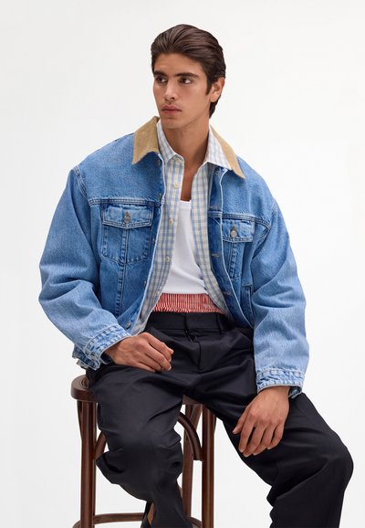 Junger Mann sitzt auf einem hölzernen Hocker, trägt eine blaue Jeansjacke, kariertes Hemd, weißes Tanktop und schwarze Hose vor weißem Hintergrund.