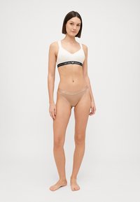 White sports bra with black logo band and light tan seamless thong with embossed branding. Both are made from soft, stretchable fabric.
