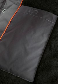 Intérieur d'une veste en polaire noire avec une poche gris foncé bordée d'orange. Texture lisse et détails de couture visibles.