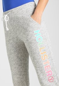 Grå sweatpants med dragsko i midjan, med en färgglad "HOLLISTER" grafik längs sidan. Mjuk, melerad textur.