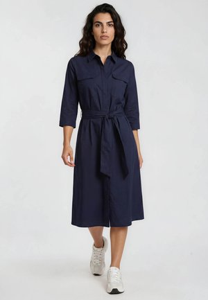 Woman walking forward wearing a navy blue belted midi dress with three-quarter sleeves and white sneakers against a plain light background.