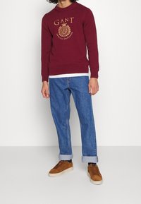 Bordeaux gebreide trui met gouden logo, gecombineerd met lichtblauwe losse jeans en bruin suède sneakers. Eenvoudig ontwerp, lange mouwen, ronde hals.