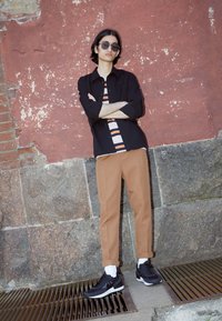 Camisa preta de botões sobre uma t-shirt riscada, calças castanhas ajustadas e ténis pretos com solas brancas. O fundo apresenta uma parede texturizada vermelha.