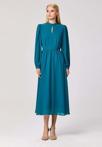 Robe longue teal à manches longues avec un col en trou de serrure, taille froncée et jupe fluide. Tissu lisse avec un léger éclat, portée avec des talons à lanières.