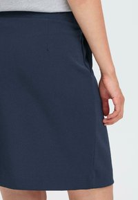 Gonna blu navy realizzata in tessuto morbido, con un design semplice, tasche laterali e orlo pulito, con una mano appoggiata sui fianchi.
