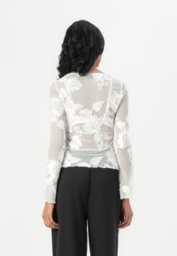 Sheer white blouse with floral embroidery, long sleeves, and a rounded hem. The fabric has a delicate texture and detailed floral patterns.