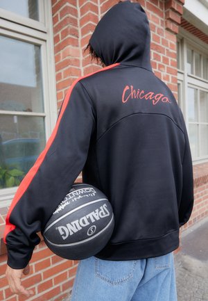 Osoba w czarnej bluzie z kapturem z napisem „Chicago” i czerwonymi paskami trzyma czarną piłkę do koszykówki marki Spalding, stojąc przy ceglanej ścianie z oknami.