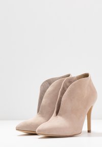 Béžové semišové kotníkové boty s špičatou špičkou, vysokým stiletto podpatkem a jedinečným výřezem podél předního otvoru. Hladký povrch po celé ploše.