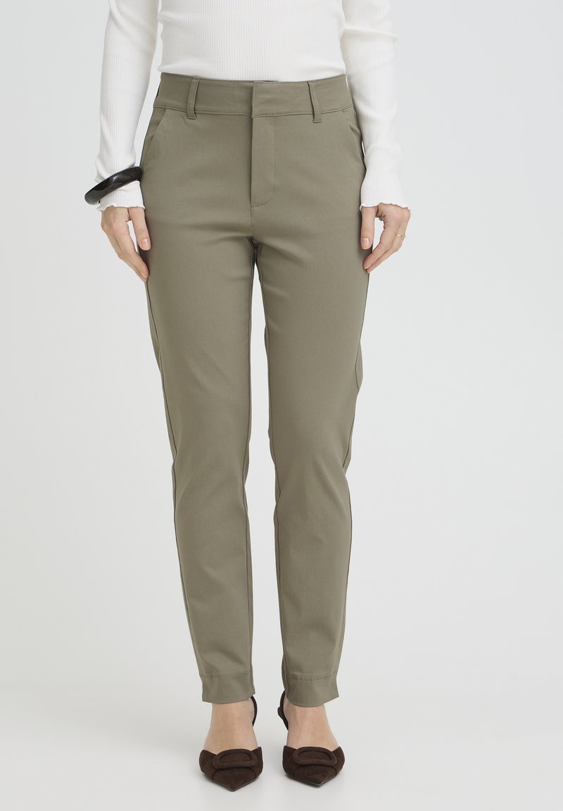 Olivgrüne, maßgeschneiderte Hose mit schmaler Passform, ausgestattet mit Seitentaschen, flacher Vorderseite und glatter Stoffstruktur. Kombiniert mit braunen spitzen Schuhen.
