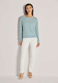 Frau, die an einer Wand steht und einen hellblauen Kabelstrickpullover, weiße Hosen und beigefarbene High-Heels trägt.