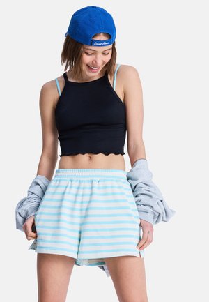 Junge Frau lächelt, trägt eine blaue Mütze rückwärts, ein schwarzes kurzgeschnittenes Tanktop, hellblau gestreifte Shorts und hält einen grauen Pullover in der Hand.