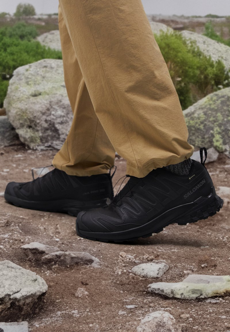 Black hiking shoes with a textured upper, lacing system, and a rugged sole, paired with loose tan pants, on rocky terrain.