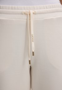 Cream-colored sweatpants with a drawstring waist, featuring gold-accented tips. The texture is smooth, and the fabric appears soft.