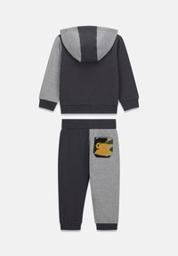 Ensemble de survêtement deux pièces en gris et bleu marine, composé d'un sweat à capuche et d'un pantalon assorti. Le pantalon présente une poche camouflage avec le logo "GUESS".
