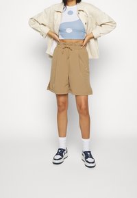 Beige shorts med resår i midjan och sidofickor, kombinerade med en blå och vit crop top med cirkulära mönster, samt vita strumpor med sportskor.