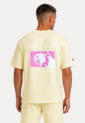 Hombre con pantalones cortos amarillo pálido y camiseta a juego con logo de felino rugiente rosa y blanco y texto en la espalda.