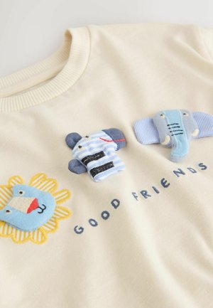 Krem pulover z tri barvnimi, teksturiranimi živalskimi aplikacijami: lev, medved v črtastem oblačilu in slon, pod njimi vezen napis.