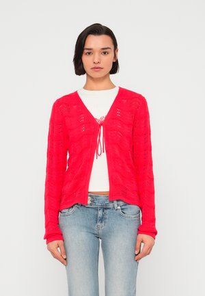 Cárdigan rojo de punto con un patrón de ondas, escote en V y detalle de lazo delantero. Mangas largas y un ajuste relajado, combinado con jeans azul claro.