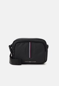 Črn najlonov crossbody nahrbtnik z zadrgo, ki ima vertikalni poudarek v rdeči, beli in modri barvi. Prikazan je logotip "TOMMY HILFIGER".