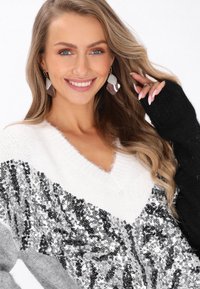 Sweter dzianinowy z dekoltem w kształcie litery V, ozdobiony srebrnymi cekinami na białym podłożu i czarnymi rękawami. Miękka faktura z luźnym krojem.