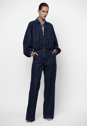 Vrouw draagt een donkerblauwe spijkerjas met elastische taille en wijde jeans, paarse sjaal, zilveren armband, staand met handen in de zakken.