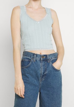 Mouwloze lichtblauwe gebreide crop top met kanten rand, gedragen met losse blauwe spijkerbroek met hoge taille, één hand in de zak, tegen een effen achtergrond.