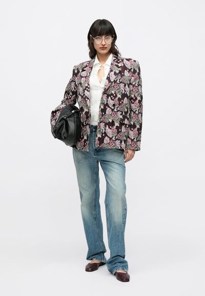 Σακάκι με floral μοτίβο σε σκούρο ύφασμα, λευκό πουκάμισο με κουμπιά, ανοιχτό μπλε τζιν παντελόνι ίσιας γραμμής και μπορντό παπούτσια, κρατώντας μια μαύρη τσάντα.