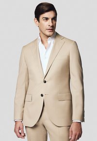 Beżowa marynarka garniturowa z dwoma czarnymi guzikami, klapami typu notch i dwiema przednimi kieszeniami. Nosi się ją na białej koszuli, dopasowanej do sylwetki.