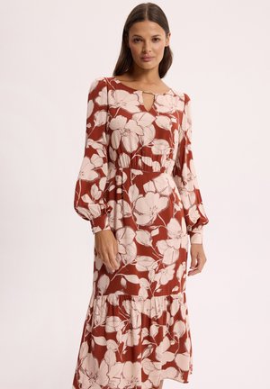 Kobieta ubrana w rdzawą sukienkę z dużym białym kwiatowym wzorem, długimi bufkami na rękawach, dekoltem z wycięciem i falbaną na dole, stojąca na tle gładkiego tła.