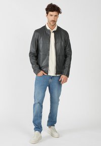 Zwarte leren bomberjack met een ronde halslijn, vooraan met rits, en zijzakken, gedragen over een crèmekleurig shirt, in combinatie met lichtblauwe jeans en witte sneakers.