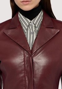 Burgundský kožený blazer s ostrými klopami, s proužkovanou košilí pod ním a knoflíky jako detaily. Hladká textura, strukturovaný design.