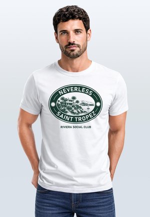Mann mit dunklem lockigem Haar und Bart, der ein weißes T-Shirt mit einem grünen "Neverless Saint Tropez" Riviera Social Club-Logo trägt.