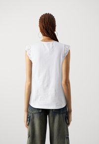 Top blanco sin mangas con detalles de encaje en los hombros. La tela es suave, con un corte holgado, que complementa los pantalones cargo de denim.