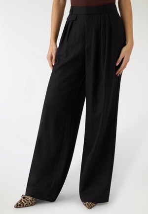 Pantaloni neri a vita alta con gamba larga e pieghe frontali, indossati con scarpe a punta stampa leopardo e una maglia marrone.